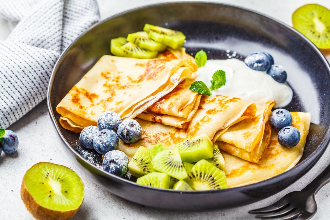 Thin crepes with curd cream, kiwi and blueberries in a black plate. Russian cuisine concept.